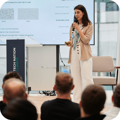 A woman speaks at a tech event, standing beside a presentation screen. Audience members are visible in the foreground.