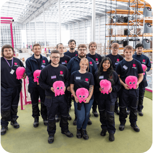 A group of people in a spacious industrial setting, each holding a pink octopus plush toy, smiling and wearing matching dark uniforms.