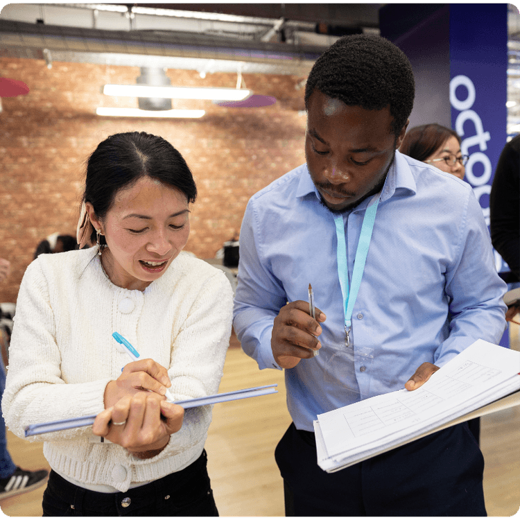A man looks over a woman's shoulder to take notes as she writes something down