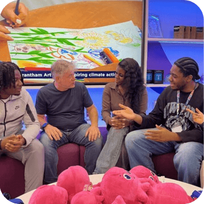 A group of people sit chatting, a colour drawing in the background and a lot of bright pink Octopus plushies in the foreground