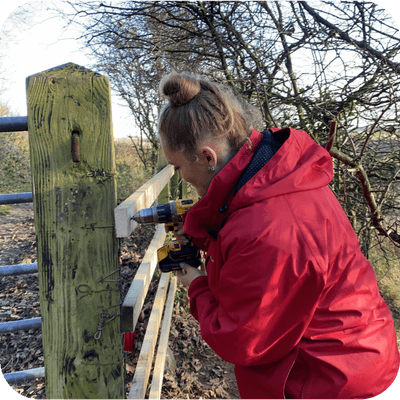 Person in a red jacket using a yellow drill to fix a wooden plank to a fence post outdoors, surrounded by trees.